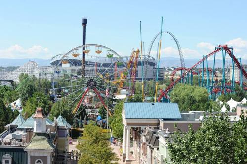 A vibrant amusement park scene featuring a Ferris wheel, roller coasters, and lush greenery under a clear blue sky.