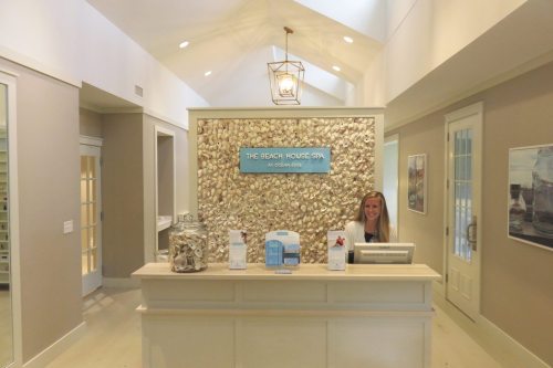 A spa reception area with a shell-covered wall and a smiling attendant at the front desk. Bright, airy atmosphere.