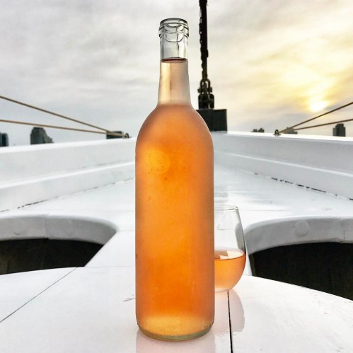 A bottle of rosé wine stands on a boat deck, with a glass of wine beside it, against a sunset backdrop.