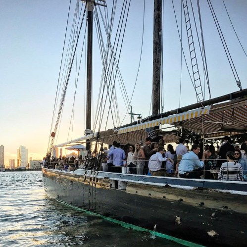 A large sailing ship with people on deck, set against a sunset skyline of tall buildings.