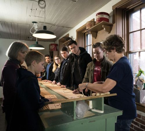 A group of people, including children and adults, gather around a table, engaged in an activity in a well-lit room.
