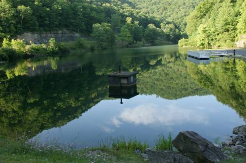 A serene lake surrounded by lush green hills, reflecting the trees and sky, with a small dock visible on the shore.