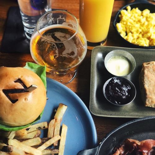 A plate with a burger, fries, scrambled eggs, and condiments, alongside a glass of beer and a drink.