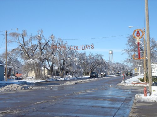 A winter street scene with icy trees, a "Happy Holidays" sign, and a water tower in the background under a clear blue sky.