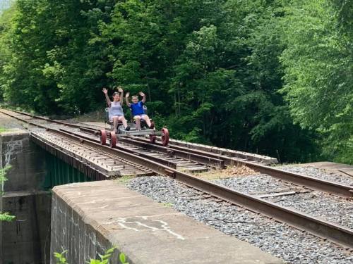 Two people ride a rail bike on train tracks surrounded by lush green trees, smiling and waving.
