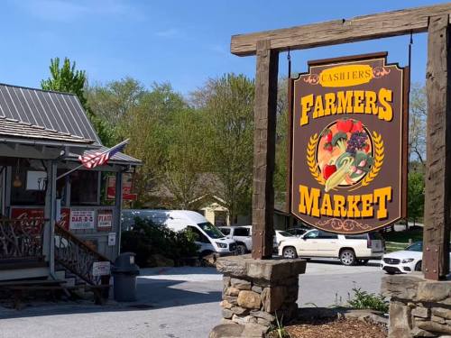 Sign for "Farmers Market" outside a rustic building, with an American flag and greenery in the background.