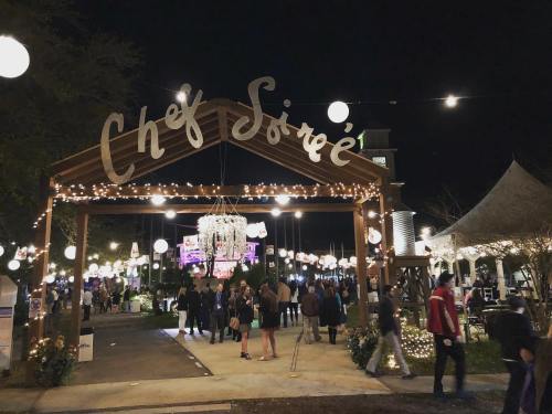 A festive outdoor event entrance at night, adorned with lights and decorations, with people mingling and enjoying the atmosphere.