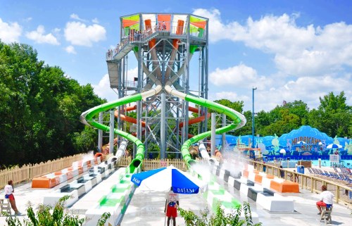 A water park featuring a tall slide tower with green and orange slides, surrounded by trees and a blue sky.