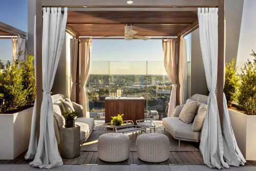Rooftop lounge area with cozy seating, a table, and plants, offering a panoramic view of the city at sunset.