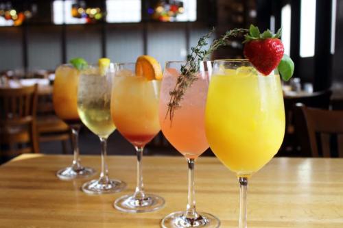 A row of colorful cocktails in tall glasses, garnished with fruit and herbs, on a wooden table in a restaurant.