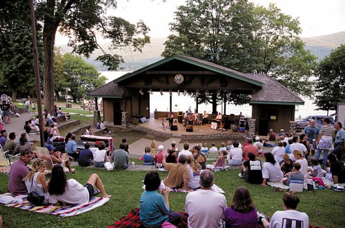 A crowd enjoys a live music performance at an outdoor stage by a lake, surrounded by trees and grassy seating areas.