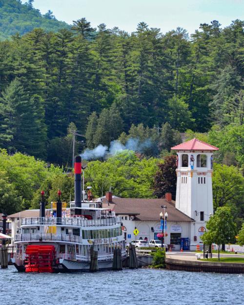 A steamboat docked by a riverside building with a clock tower, surrounded by lush green trees and a clear blue sky.