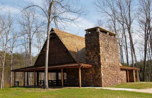 A rustic stone and wood pavilion surrounded by trees, featuring a tall chimney and picnic tables underneath.