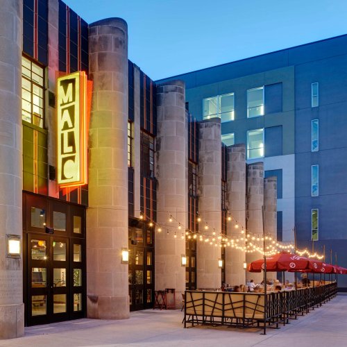 Exterior view of a building with a neon sign reading "MALC," featuring outdoor seating and string lights at dusk.