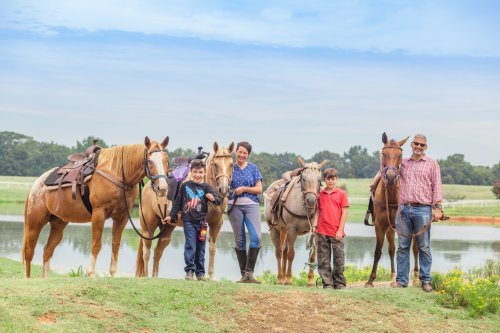 A family stands by a pond with four horses, smiling and dressed for riding, under a clear blue sky.