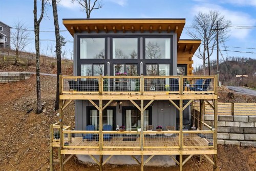 Modern two-story house with large windows and a wooden deck, surrounded by trees and a sloped landscape.