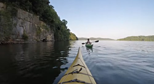 A kayaker paddles on a calm lake, surrounded by cliffs and lush greenery under a clear sky.