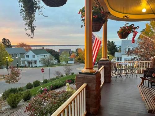 View from a porch with hanging plants, an American flag, and a scenic landscape of houses and a lake at sunset.
