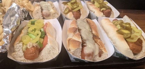 A variety of hot dogs in paper trays, topped with jalapeños, cheese, and green onions, served with curly fries.