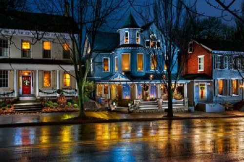 A charming street scene at night, featuring illuminated houses and wet pavement reflecting colorful lights.