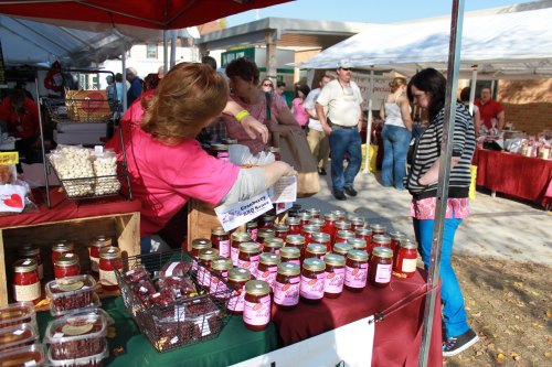 A bustling outdoor market with vendors selling jars of jam and visitors browsing the stalls under colorful tents.