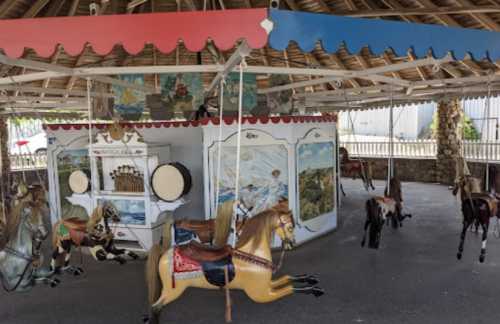 A vintage carousel with painted wooden horses, set under a thatched roof in a sunny outdoor area.