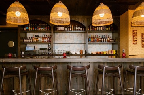 A cozy bar with wooden shelves stocked with bottles, illuminated by woven pendant lights and surrounded by high stools.