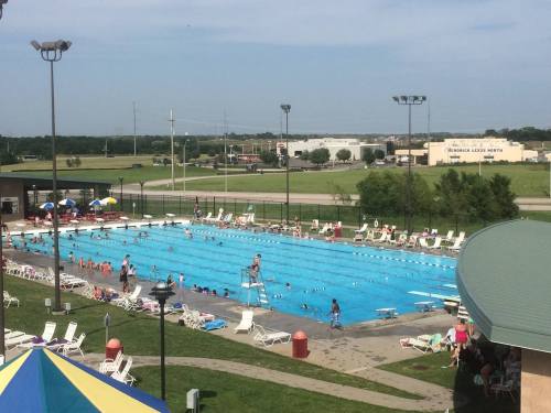 A large outdoor swimming pool filled with people, surrounded by lounge chairs and umbrellas on a sunny day.