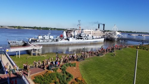 A large ship docked by a river, with a crowd of people gathered on the shore and clear blue skies above.