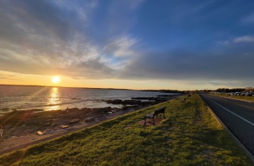 A serene sunset over a calm sea, with a grassy shoreline and benches along a winding road.