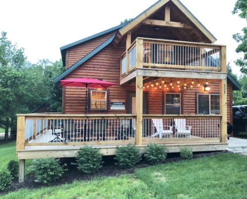 A cozy log cabin with a wooden deck, patio furniture, and string lights, surrounded by greenery.