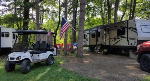 A campsite with a golf cart, American flag, and a parked RV surrounded by trees and outdoor seating.