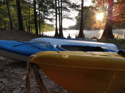Two kayaks, one blue and one yellow, are resting on a shore with trees and a misty river in the background at sunset.