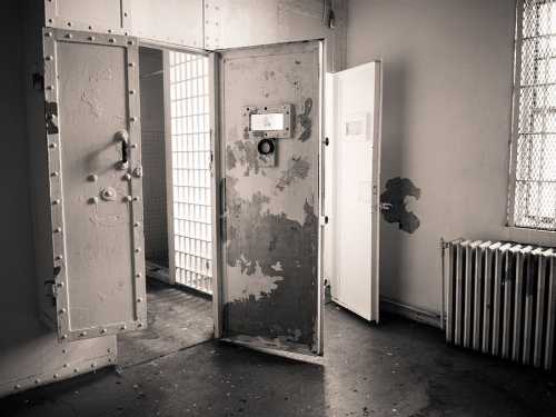 An empty, dimly lit prison cell with open metal doors and a radiator, showcasing peeling paint and barred windows.