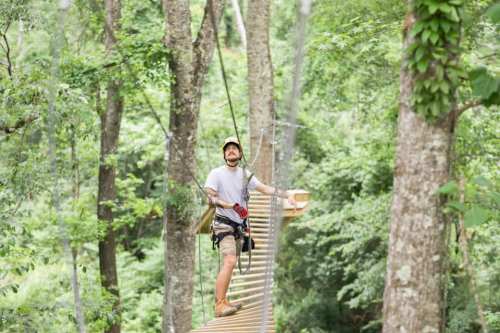 A person on a rope bridge in a lush green forest, smiling and wearing safety gear.