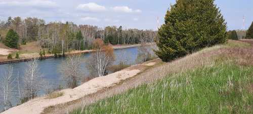 A serene river flows beside grassy banks and trees, under a partly cloudy sky.