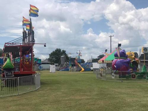 A colorful carnival scene with rides, games, and flags under a partly cloudy sky. Green grass in the foreground.