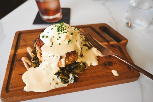 A wooden plate holds a crab cake topped with creamy sauce and chives, served with greens and a fork beside it.