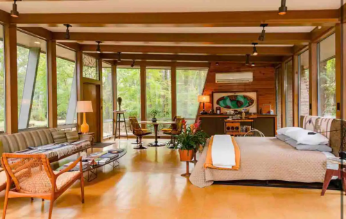 A modern, airy room with large windows, a bed, seating area, and stylish decor surrounded by greenery.