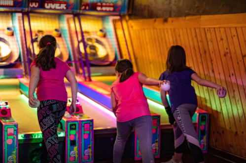 Three girls playing a skee-ball game in a colorful arcade, focused on their throws.