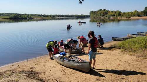 A group of people preparing canoes on a sandy riverbank, with others paddling in the background on a sunny day.