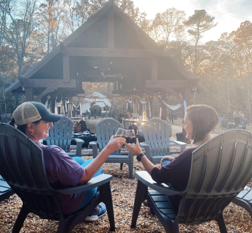 Two people sitting in adirondack chairs, toasting drinks in a cozy outdoor setting with trees and a rustic building in the background.