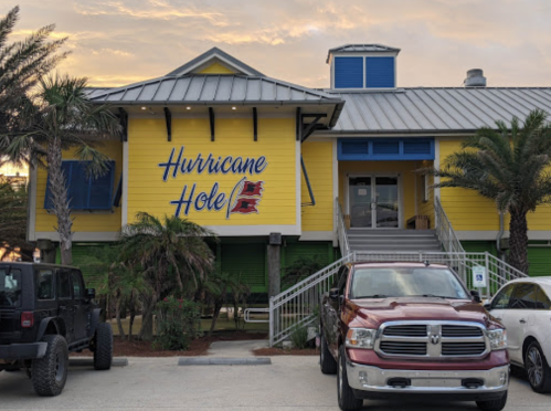 A colorful building with "Hurricane Hole" sign, surrounded by palm trees and parked vehicles under a sunset sky.
