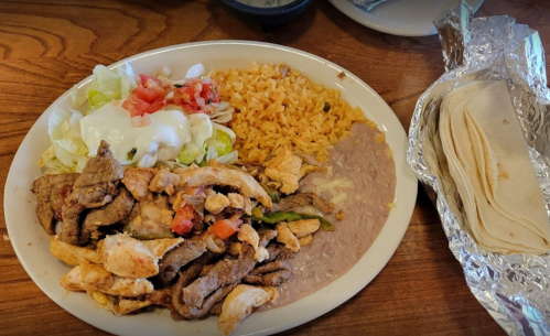 A plate of grilled chicken and beef with rice, refried beans, lettuce, tomatoes, sour cream, and a side of tortillas.