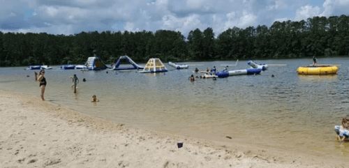 A sandy beach with people swimming and playing on inflatable water structures in a lake surrounded by trees.