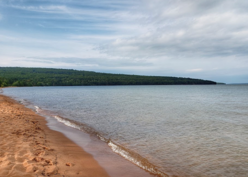 A serene beach scene with gentle waves lapping at the sandy shore, surrounded by lush green hills under a cloudy sky.