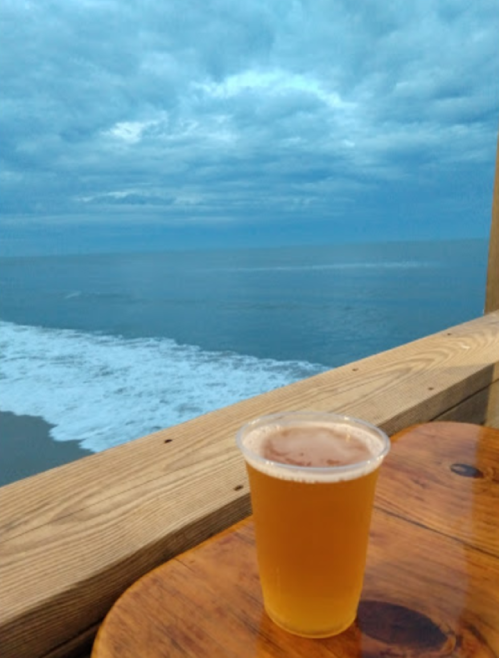 A cup of beer sits on a wooden table overlooking a calm ocean under a cloudy sky.