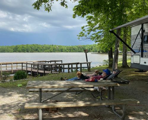 A peaceful lakeside scene with two people relaxing at a picnic table, a dog nearby, and an RV in the background.