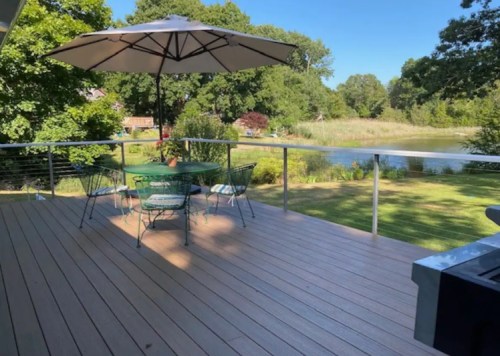 A serene deck with a table and chairs under an umbrella, overlooking a pond and lush greenery.