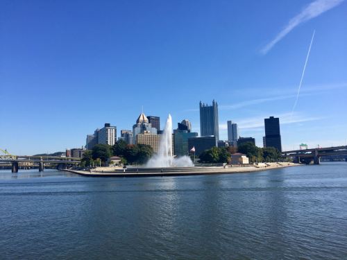 City skyline of Pittsburgh with a fountain in the foreground, clear blue sky, and calm river.
